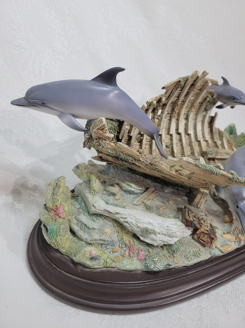 May include: A figurine of three dolphins jumping out of the water near a shipwreck. The dolphins are grey and white, and the shipwreck is made of wood. The figurine is set on a base that is covered in green moss and has a treasure chest.