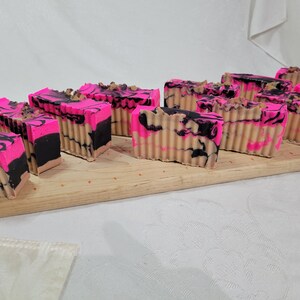 May include: A close-up of a wooden board with multiple bars of handmade soap. The soap is pink, black, and beige with a marbled swirl pattern.