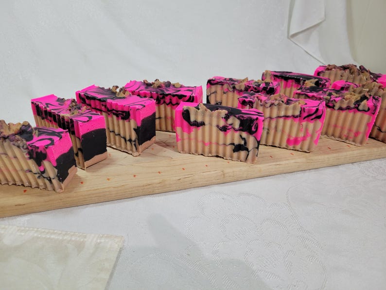May include: A close-up of a wooden board with multiple bars of handmade soap. The soap is pink, black, and beige with a marbled swirl pattern.