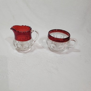 Puede incluir: Un juego de jarra de crema y azucarero de cristal transparente con borde y detalles en rojo rubí. La jarra de crema tiene un asa y una parte superior con volantes. El azucarero tiene un asa y un diseño circular. Ambas piezas están sobre una superficie blanca.