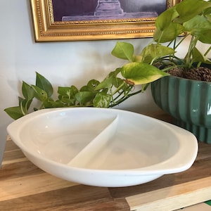Puede incluir: Un plato de servir ovalado blanco dividido con un asa a cada lado. El plato está sobre una tabla de cortar de madera. Una planta verde en una maceta verde azulado está en el fondo, junto con un cuadro enmarcado.