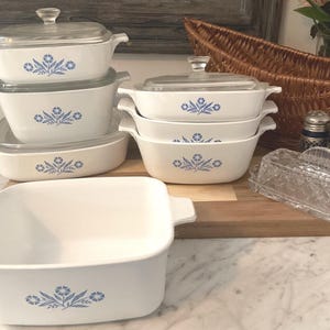 Könnte beinhalten: Eine Sammlung weißer Keramik-Auflaufformen mit blauen Blumenmustern und Glasdeckeln. Das Set umfasst verschiedene Größen und Formen, ideal zum Backen und Servieren. Eine klare Glasbutterdose und Salz- und Pfefferstreuer sind ebenfalls vorhanden.