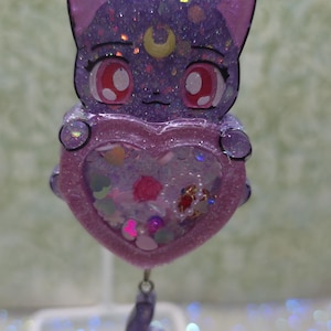 May include: A purple cat-shaped keychain with a heart-shaped shaker filled with colorful glitter and charms. The keychain has a silver clasp and a small, dangling tail charm. The cat has large red eyes and a crescent moon on its forehead.