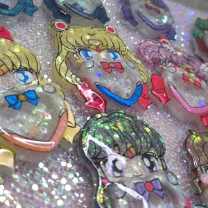 May include: A collection of glittery resin charms featuring anime-style characters with large eyes, bows, and colorful hair. The charms are set against a sparkling, iridescent background, creating a whimsical and decorative effect.