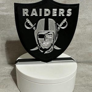 May include: A black and silver Raiders logo, featuring the team's shield emblem with the word "RAIDERS" in silver lettering. The logo includes a silver helmet and crossed swords. The logo is mounted on a white base.