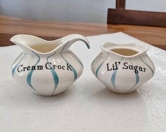 May include: White ceramic creamer and sugar set with blue striped designs. The creamer is labeled "Cream Crock" and the sugar bowl is labeled "Lil' Sugar".