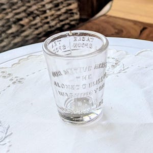 May include: A clear, vintage shot glass with etched text. The glass reads "OUR NATIVE HELLS THE ALONZO O BLISS WASHINGTON D.C." and has additional text at the top. The glass is on a white, embroidered doily.