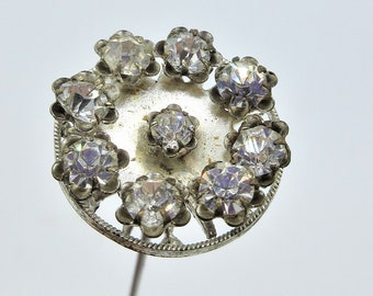 May include: A silver hat pin with a circular rhinestone design. The pin is held in a hand.