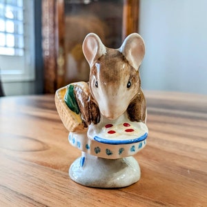 May include: A ceramic figurine of a brown mouse holding a blue and white plate with red dots. The mouse is wearing a green and brown hat and is standing on a blue and white base.