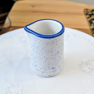 May include: A small ceramic creamer pitcher with a white base featuring a blue speckled pattern and a solid blue rim. The pitcher has a simple cylindrical body with a small pouring spout at the top, designed for serving cream or milk at the table.