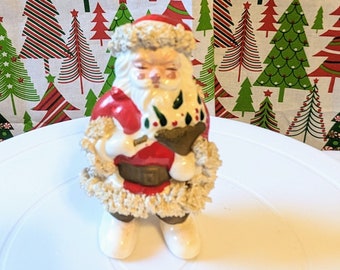 May include: A vintage ceramic Santa Claus figurine with a white beard and a red and white suit. He is holding a bag of gifts and is standing on a white surface.
