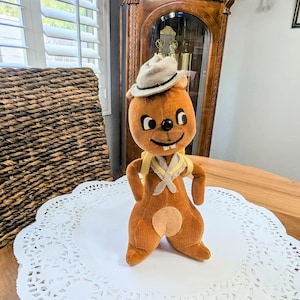 May include: A vintage plush bear toy with a small hat and yellow scarf, standing on a white paper doily. The bear is brown with a light brown patch on its belly. The background includes a wooden table and a grandfather clock.