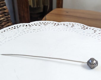 May include: A silver pin with a decorative round head on a white doily.