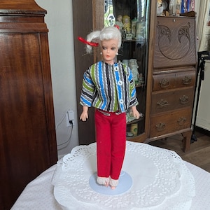 May include: A vintage doll with white hair styled in a ponytail, wearing a striped jacket in blue, green, and black, paired with red pants. The doll is standing on a white lace doily, showcasing a retro fashion aesthetic.
