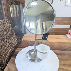 May include: A vintage shaving set featuring a round, silver-framed mirror, a shaving brush, and a white ceramic cup with a silver rim. The set is mounted on a white, round base with a lace doily. The background includes a wooden table and a grandfather clock.