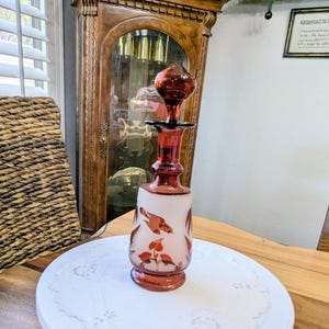 May include: A vintage red glass decanter with a matching stopper. The decanter features a white frosted body with painted bird and floral designs. It is displayed on a white cake stand with a decorative paper doily.