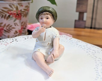 May include: A porcelain figurine of a seated baby wearing a white dress with gold accents and a green hat. The baby is posed with one hand near its mouth and the other resting on its foot. The figurine is on a white doily.