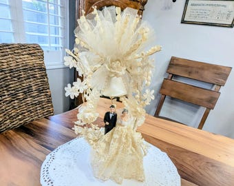 May include: A vintage wedding cake topper featuring a bride and groom figurine. The topper is adorned with cream-colored lace, flowers, and a bell. It rests on a white paper doily, creating a classic and romantic aesthetic.