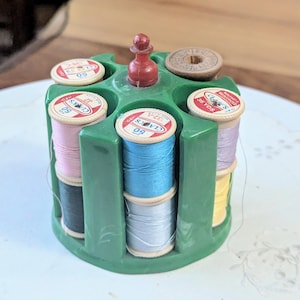May include: A green plastic circular thread spool holder with a central red knob, containing multiple spools of Clark's brand sewing thread in various colors including pink, blue, yellow, and black, with one empty wooden spool visible on top.