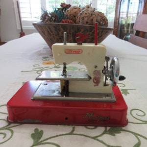 May include: A red and white toy sewing machine with a silver needle and a black foot pedal. The machine is labeled "Jet Sew-Matic" and "Elraco".