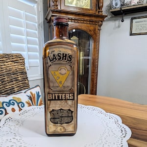 May include: A vintage glass bottle of Lash's Bitters. The bottle is brown with a yellow label that reads "Lash's Bitters" and "Return Label". The label also has a yellow triangle with the letters "L.P.C." inside. The bottle is sitting on a white doily.