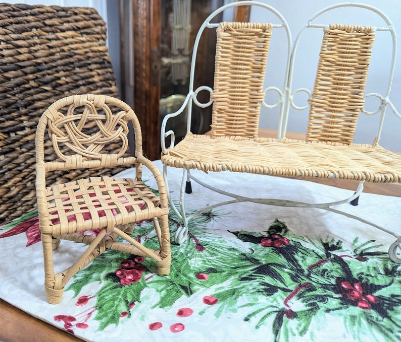 May include: A miniature wicker chair with a woven back and seat, next to a small white metal and wicker bench. The chair has a decorative knot design on the back. The bench has a curved metal frame. The items are on a festive tablecloth.