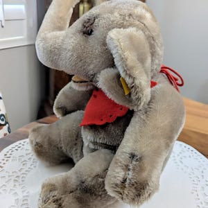 Pode incluir: Um brinquedo de elefante de pel&uacute;cia cinza com uma bandana vermelha em volta do pesco&ccedil;o. O elefante tem uma tromba longa e orelhas grandes. O brinquedo est&aacute; sentado em um croch&ecirc; branco.