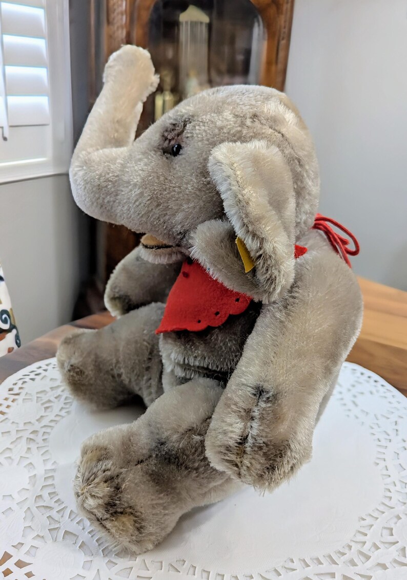 Pode incluir: Um brinquedo de elefante de pel&uacute;cia cinza com uma bandana vermelha em volta do pesco&ccedil;o. O elefante tem uma tromba longa e orelhas grandes. O brinquedo est&aacute; sentado em um croch&ecirc; branco.
