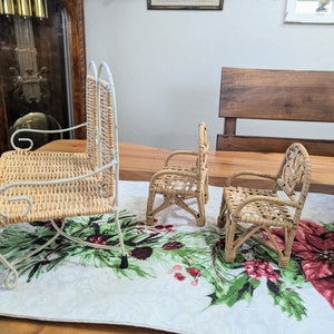 May include: Three miniature wicker chairs. One is a rocking chair with a white metal frame. The other two are armchairs. All three chairs are made of woven light brown wicker. The chairs are on a table with a Christmas-themed runner.