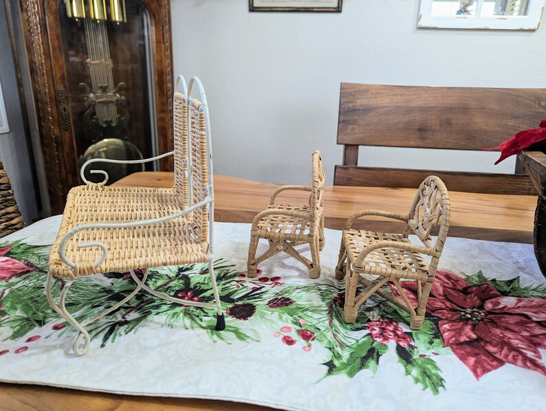 May include: Three miniature wicker chairs. One is a rocking chair with a white metal frame. The other two are armchairs. All three chairs are made of woven light brown wicker. The chairs are on a table with a Christmas-themed runner.