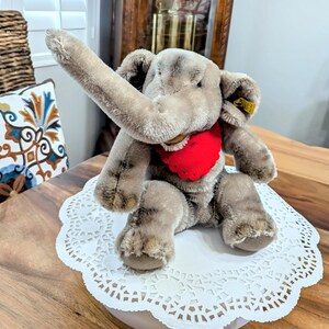 Pode incluir: Um brinquedo de elefante de pel&uacute;cia cinza com um babador vermelho em forma de cora&ccedil;&atilde;o, sentado em um la&ccedil;o branco. O elefante tem uma tromba longa levantada no ar e um bot&atilde;o Steiff na orelha.