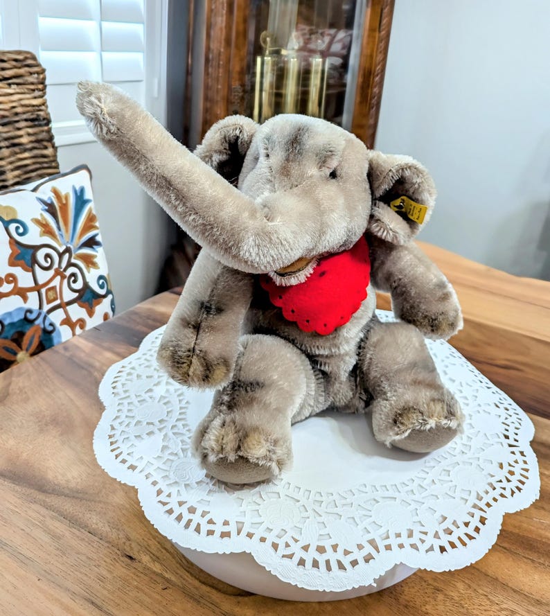 Pode incluir: Um brinquedo de elefante de pel&uacute;cia cinza com um babador vermelho em forma de cora&ccedil;&atilde;o, sentado em um la&ccedil;o branco. O elefante tem uma tromba longa levantada no ar e um bot&atilde;o Steiff na orelha.
