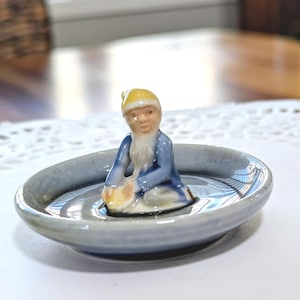 May include: A small, ceramic figurine of a seated man in a blue robe and yellow hat, placed in a gray, shallow dish. The figurine has a white beard and is holding a yellow object. The dish has a reflective surface.