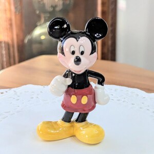 May include: A ceramic Mickey Mouse figurine. The figure has black ears, a black body, red shorts with yellow buttons, white gloves, and yellow shoes. The figurine is standing on a white doily.