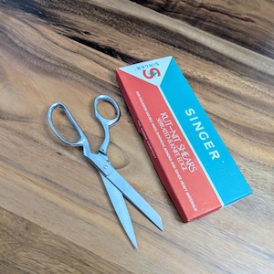 May include: A pair of silver Singer Kut-Nit shears with serrated and knife edges. The shears are packaged in a blue and red box with the Singer logo and the text "Kut-Nit Shears, Serrated & Knife Edge, For Polyester Double Knits, Synthetic Bonded and Other Heavy Materials."