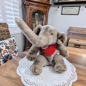 Pode incluir: Um brinquedo de elefante de pel&uacute;cia cinza com uma bandana vermelha. O elefante tem uma tromba longa e orelhas grandes. O brinquedo est&aacute; sentado em um la&ccedil;o branco.