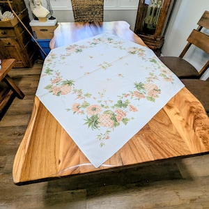 May include: A white square tablecloth with a floral and pineapple design in shades of pink, green, and yellow. The tablecloth is placed on a large wooden table. The table is surrounded by wooden chairs and a grandfather clock.