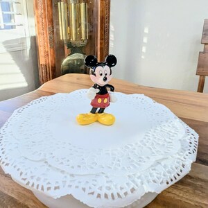 May include: A small Mickey Mouse figurine with black ears, a black body, red shorts, white gloves, and yellow shoes. The figurine is on a white paper doily. The background includes a wooden table and a grandfather clock.
