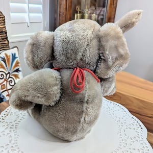 Pode incluir: Um brinquedo de elefante de pel&uacute;cia cinza com uma fita vermelha amarrada ao redor do pesco&ccedil;o. O elefante est&aacute; sentado em um la&ccedil;o branco.