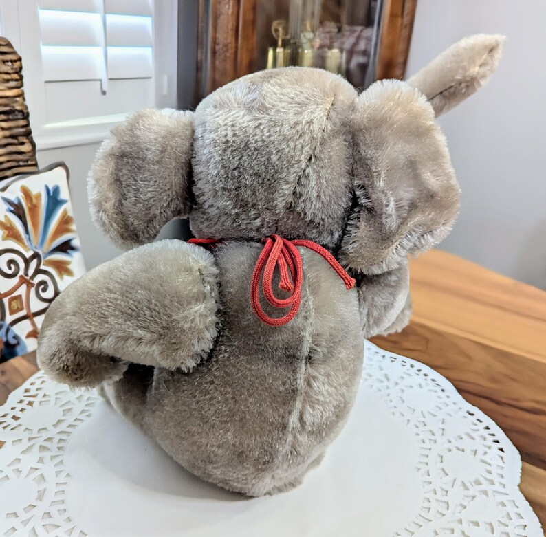 Pode incluir: Um brinquedo de elefante de pel&uacute;cia cinza com uma fita vermelha amarrada ao redor do pesco&ccedil;o. O elefante est&aacute; sentado em um la&ccedil;o branco.