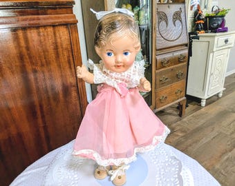 Pode incluir: Uma boneca vintage com olhos azuis, vestida com um vestido rosa com gola e acabamento em renda branca. A boneca tem um laço branco no cabelo e está em cima de uma toalha de mesa branca. Os sapatos da boneca são castanhos.
