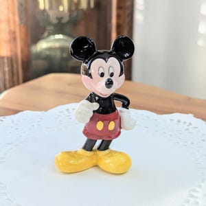 May include: A ceramic Mickey Mouse figurine. The figure has black ears, a black body, and red shorts with two yellow buttons. Mickey's shoes are yellow, and his gloves are white. The figurine is standing on a white doily.