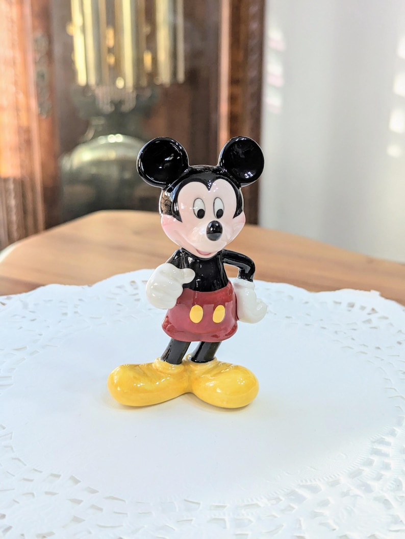 May include: A ceramic Mickey Mouse figurine. The figure has black ears, a black body, and red shorts with two yellow buttons. Mickey's shoes are yellow, and his gloves are white. The figurine is standing on a white doily.