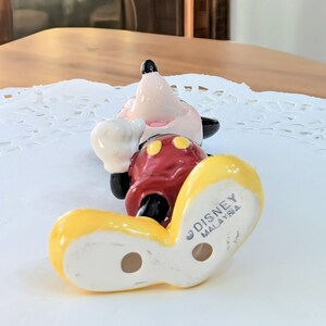 May include: A ceramic Mickey Mouse figurine in a playful pose. The figurine features a red body, yellow shoes, white gloves, and a pink face with black ears. The bottom of the shoe is stamped with "DISNEY MALAYSIA".