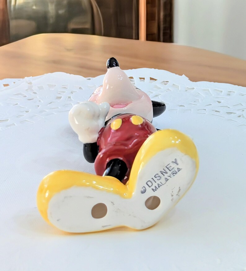May include: A ceramic Mickey Mouse figurine in a playful pose. The figurine features a red body, yellow shoes, white gloves, and a pink face with black ears. The bottom of the shoe is stamped with "DISNEY MALAYSIA".
