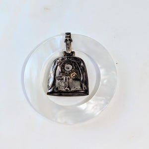 May include: A vintage silver-toned bell pendant with the text "MONTEVER" and the number "755" is suspended from a mother-of-pearl ring. The bell has a small loop at the top for hanging. The ring is white and circular.