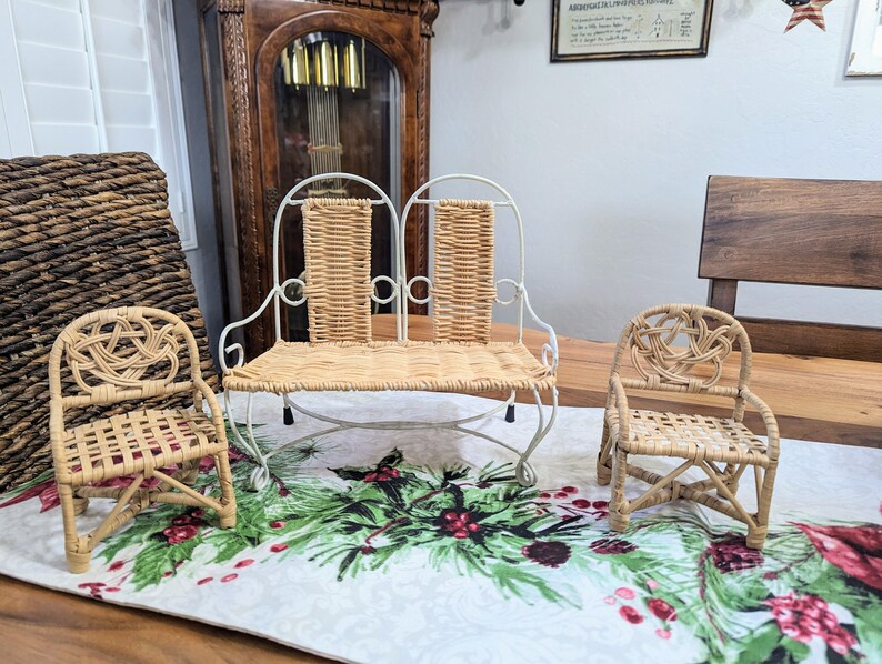 May include: Three miniature wicker chairs. A double seat bench with a white metal frame and woven back and seat. Two single chairs with woven seats and backs. The chairs are on a table runner with a Christmas theme.