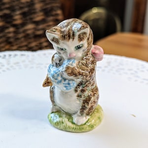 May include: A brown and white ceramic figurine of a cat holding a blue and white checkered mouse. The cat has green eyes and a pink bow. The figurine is standing on a green base.