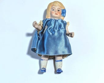 May include: A small porcelain doll with blonde hair and a blue dress. The doll is wearing white socks and blue shoes.