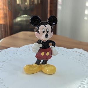 May include: A ceramic Mickey Mouse figurine. The figure has black ears, a black face, and a red shorts with two yellow buttons. The figurine is wearing yellow shoes and white gloves. The figurine is standing on a white paper doily.
